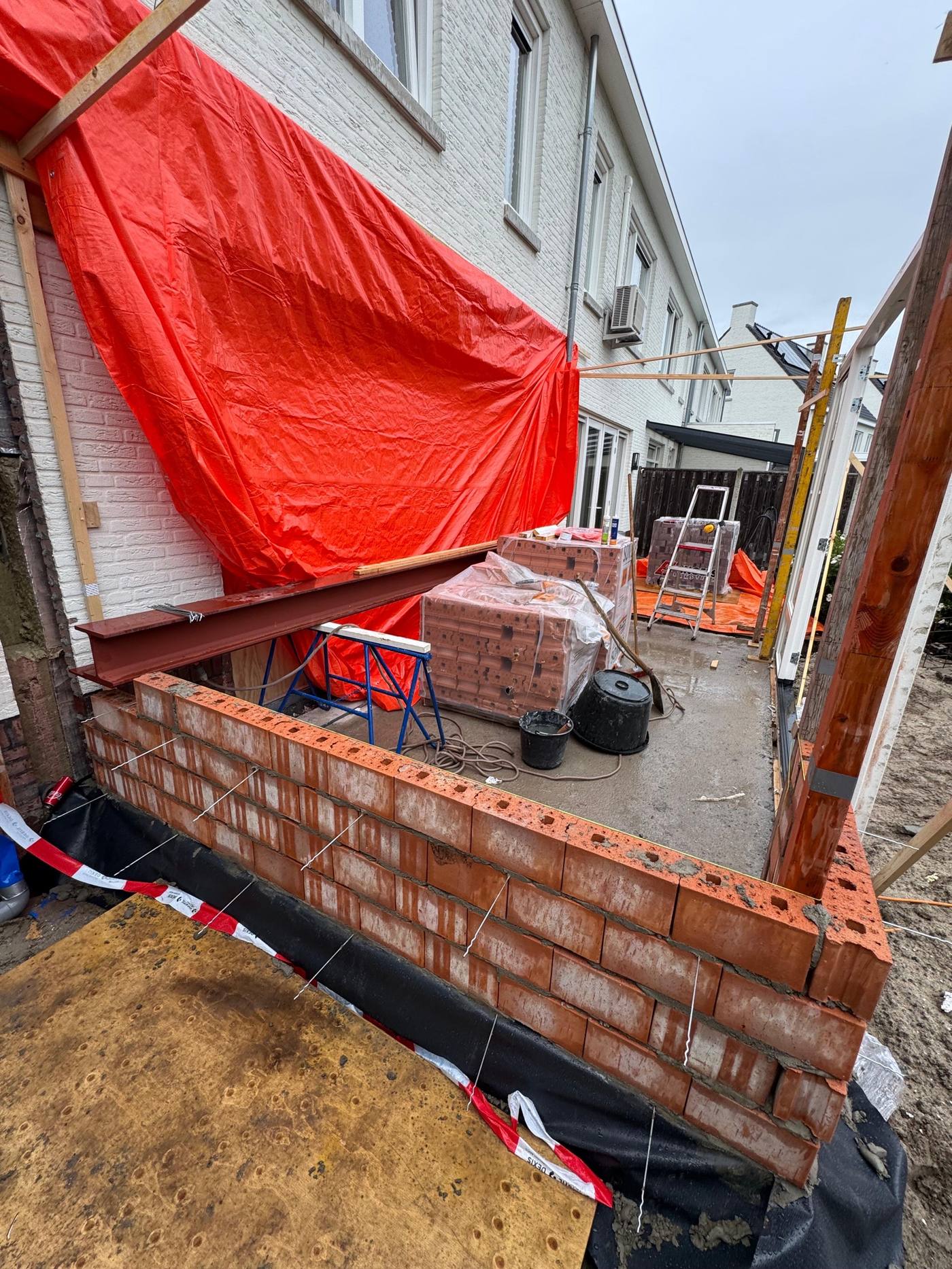 Metselwerk van de nieuwe aanbouw in opbouw, met een stalen lateibalk en pallets baksteen klaar voor verwerking